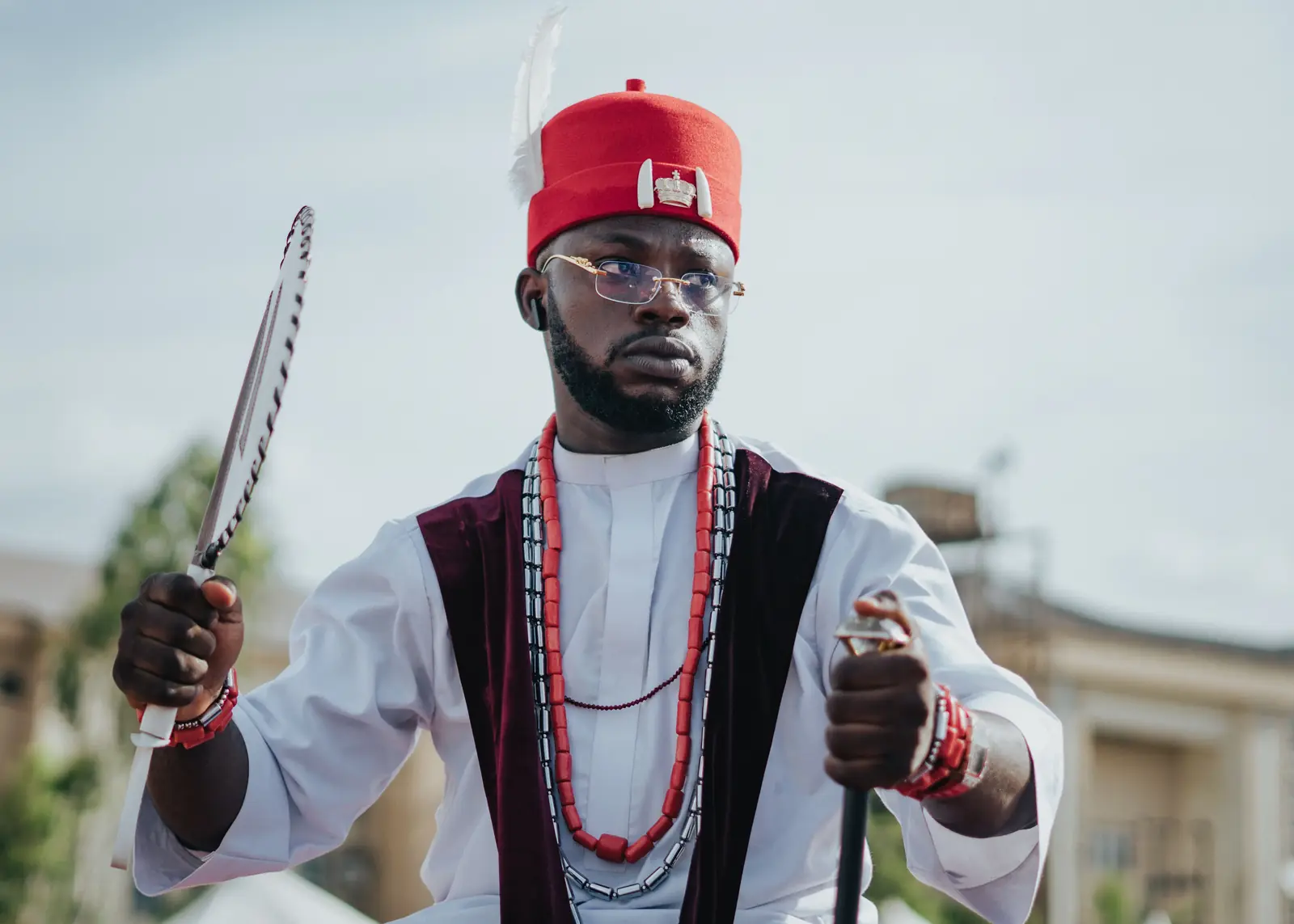A man in white with a red cap, coral beads, a hand fan and a staff